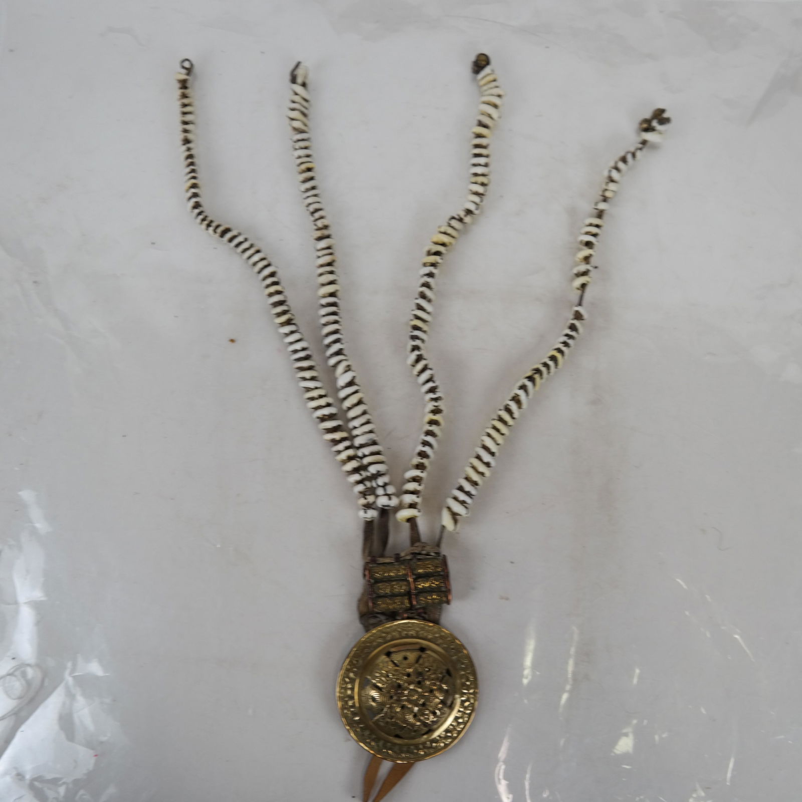 Tibetan Woman: Tibetan pendant, chabchab, having an"Endless Knot" pattern brass disc above three incised brass tube spacers, from which hang three strands of cowrie shells. [Disc: 3 5/8 (dia) inches; Overall length: