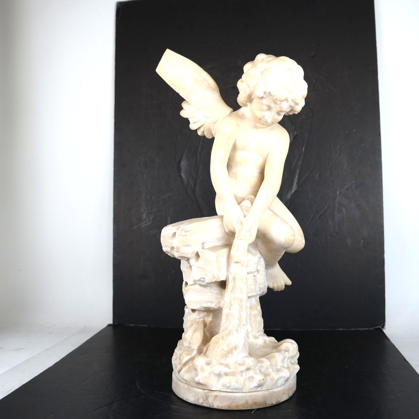 Carved Alabaster Cherub Sculpture (As-Is): Carved alabaster sculpture of a cherub on a rock, looking down. Psyche styling. [19" H x 9" Diam.]. Wings as-is (as shown).