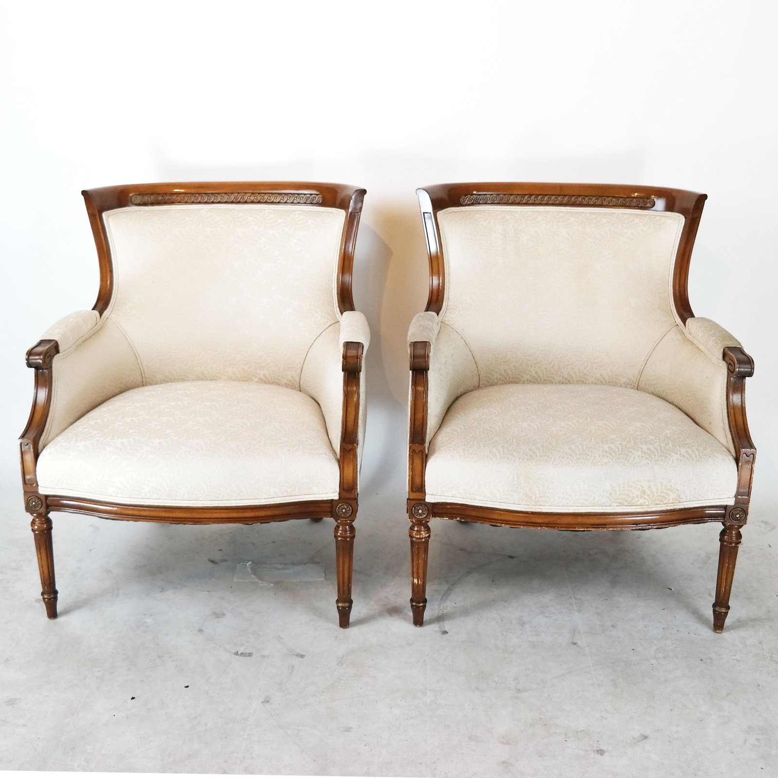 Pair of Mahogany Louis XVI-Style Bergeres: Pair of 20th century Louis XVI-style bergeres, with cream jacquard upholstery. [32" H x 26 1/2" W x 24" D]. Wear to legs.