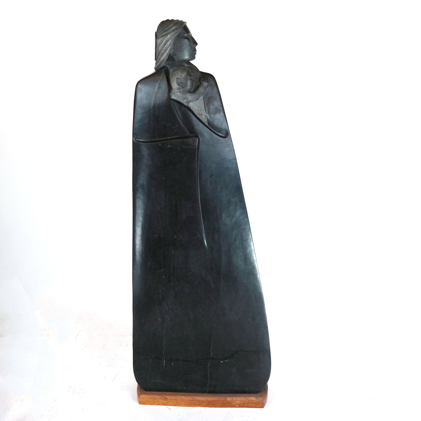 Tall Inuit-Style Stone Sculpture of a Woman & Child: Inuit-style stone sculpture of a woman holding a baby, on a short wooden base. No signature found. [Overall: 55" H x 21" W x 8" D]. Few marks and scratches.