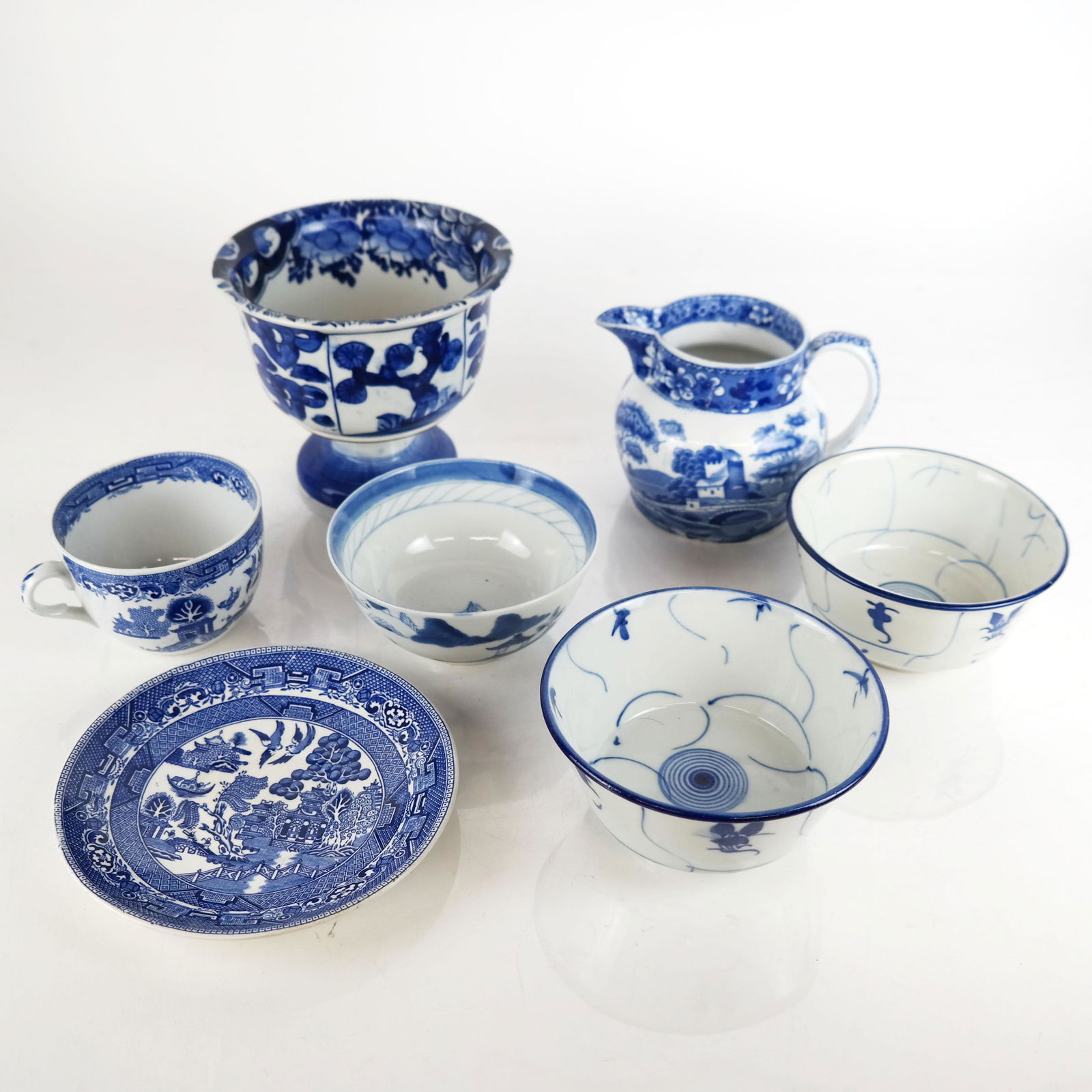 Lot of Blue & White Porcelain Table Items: Lot includes an English Blue Willow pattern cup and saucer, 3 Japanese bowls, an unmarked pedestal bowl with Japanese-style decoration, and a Spode pitcher. [Largest: 5" H x 6" Diam.]. In good conditi