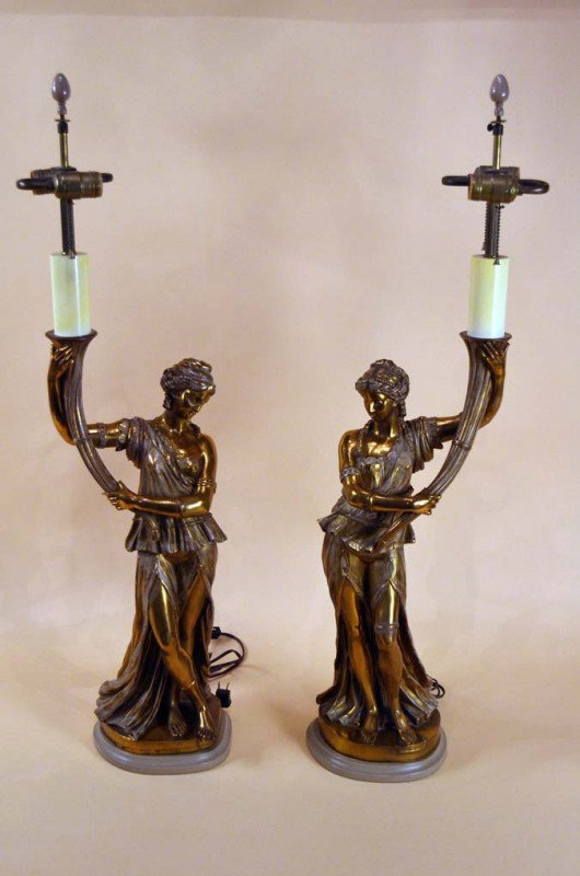 Pair of Bronze Figural Lamps: Having a pair of classical women facing one another, each holding a torch, one having crossed legs. each on wood plynth with shades. 23.5"