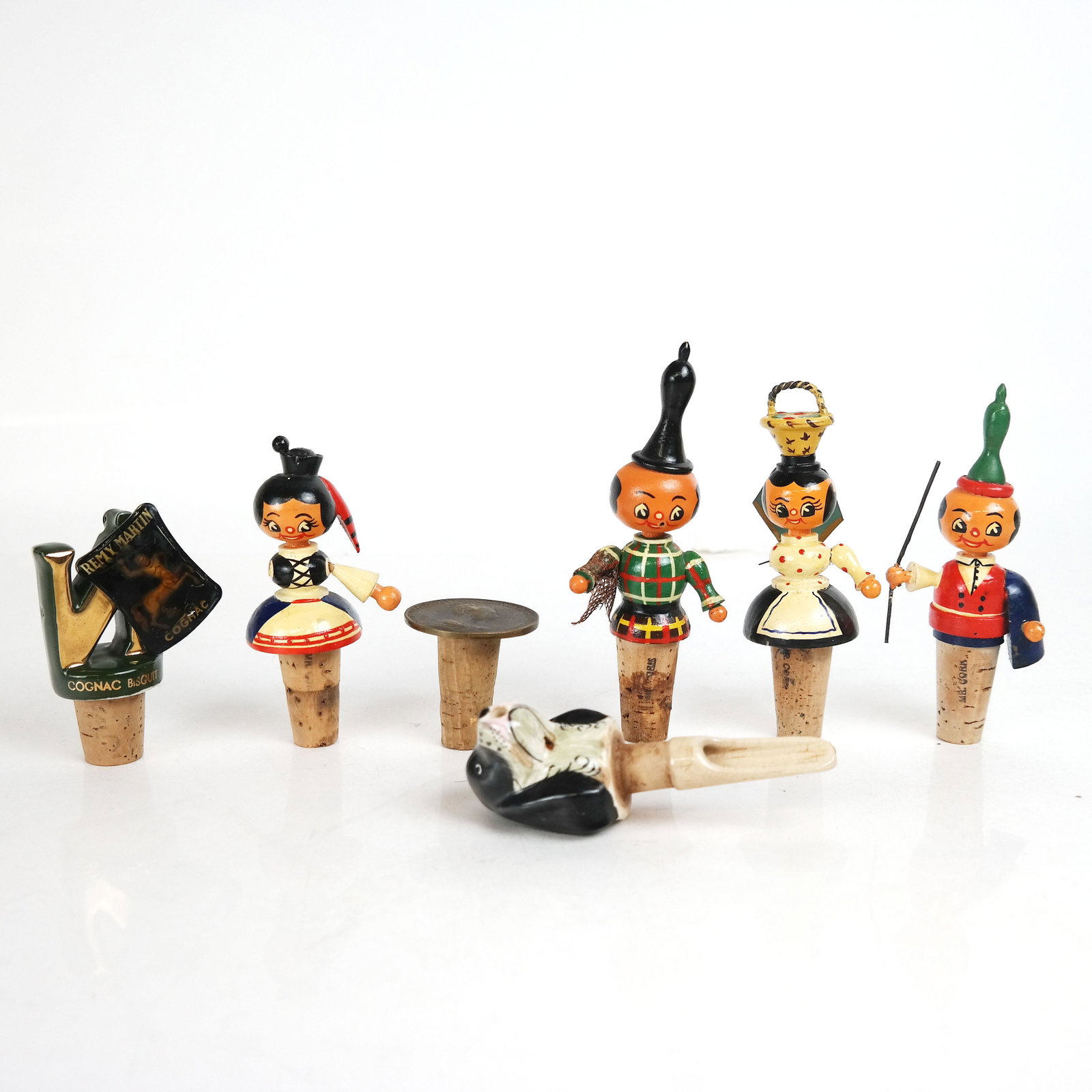 Lot of 7 Decorative Wine Stoppers: Lot of 7 decorative wine stoppers, including Remy Martin, Ceramic Staffordshire dog head, a Queen Anne coin and 4 painted dolls. From the Estate of Florence and Herbert Irving. [Tallest: 1 1/2" H]. In