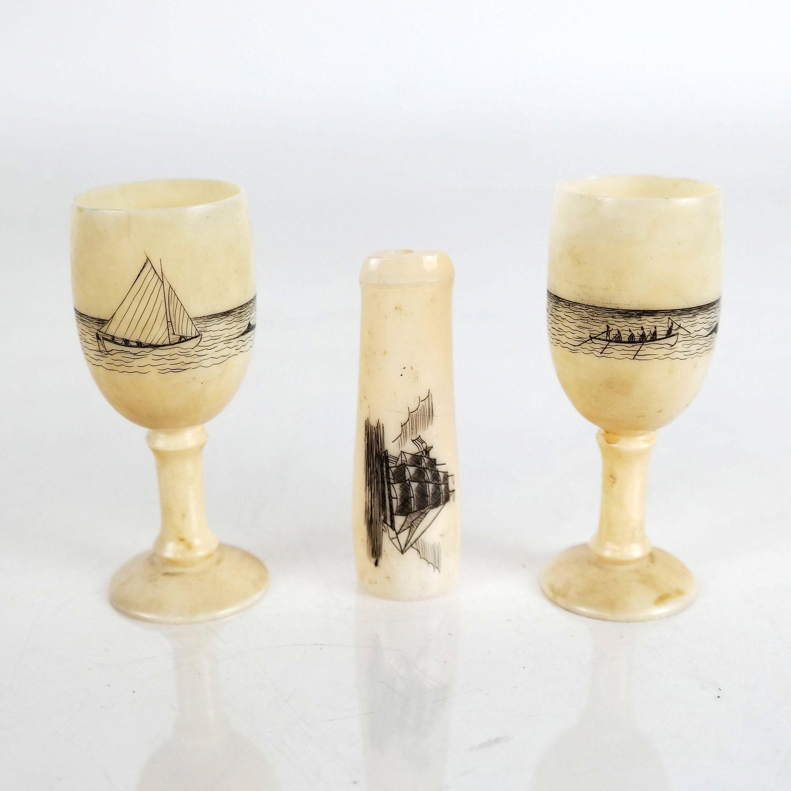 Three Antique Engraved Scrimshaw Sculptures: Lot of three 19th century American engraved sperm whale teeth, including a pair of cups with engraved nautical scenes [2 1/2" H], and a cigar tip holder [2 1/4" Long]. In good condition.