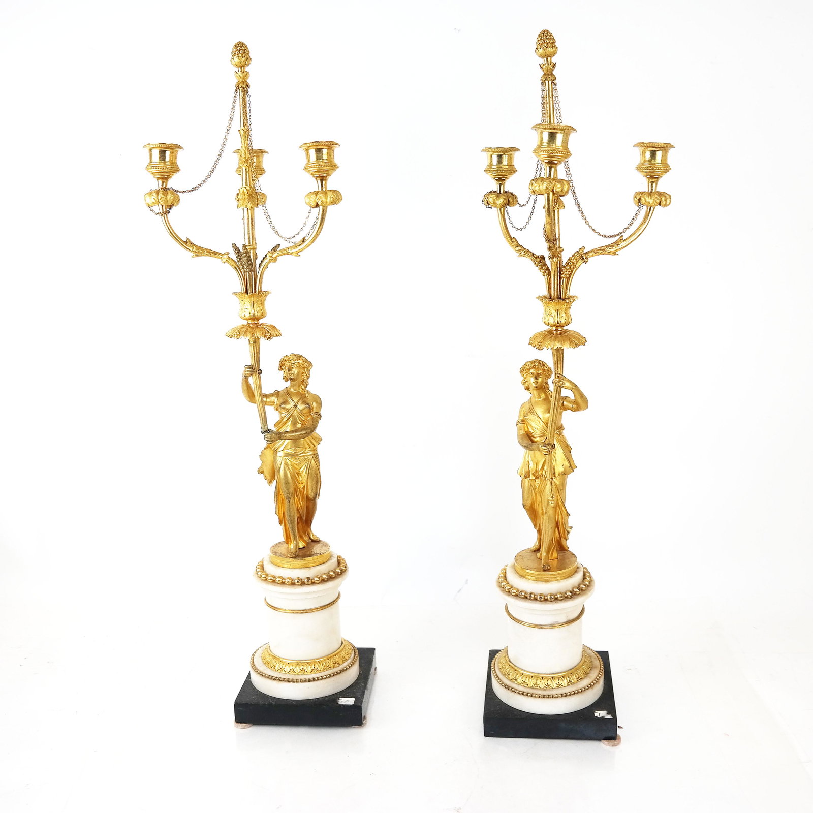 Pair of Neoclassical-Style Gilt Bronze Candelabra: The figural standards holding up cornucopia-form three-branch candle cups accented with acanthus leaves and feather plumes, the columns with bead and foliate motifs, raised on marble column and square