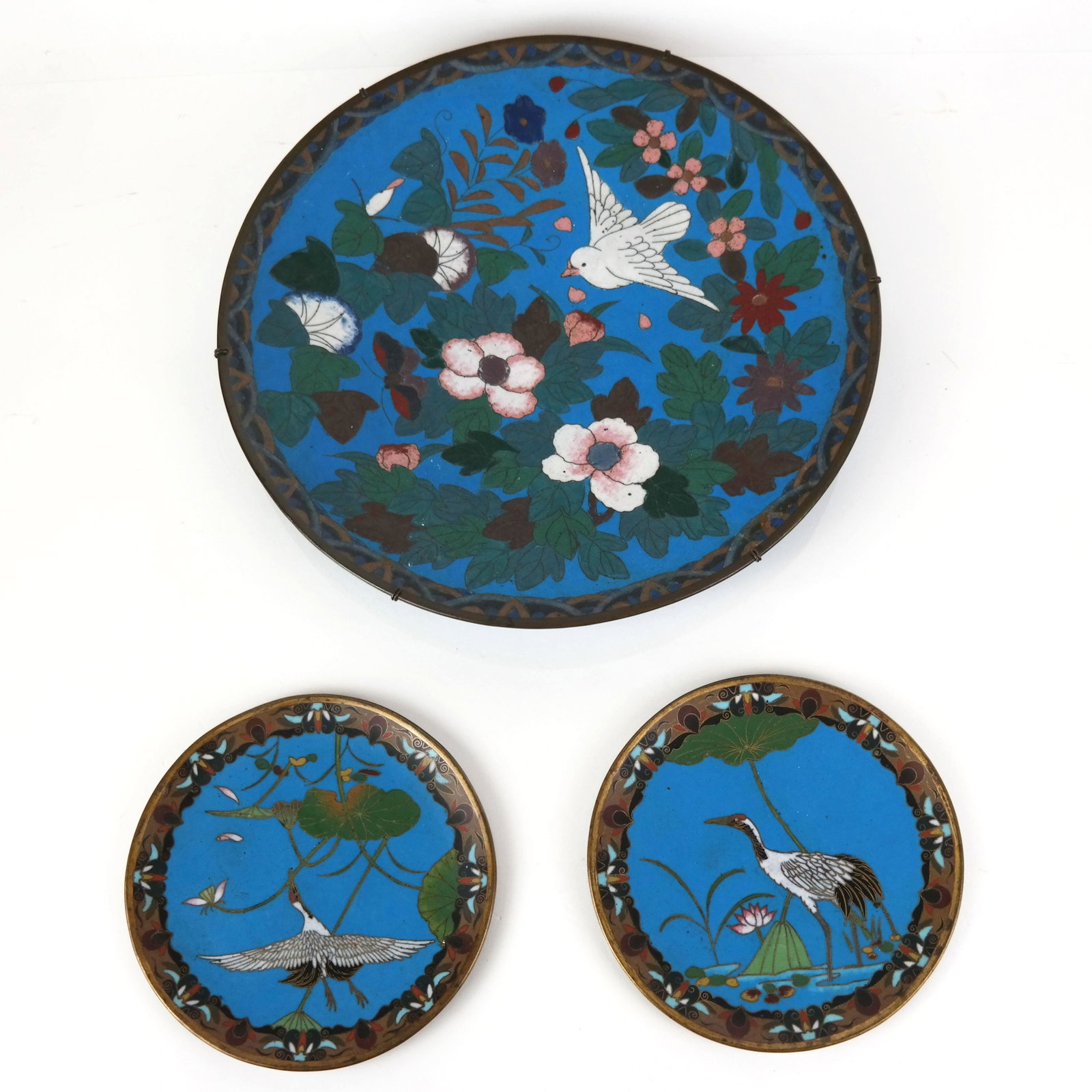 Three Japanese Cloisonne Enamel Plates: Three Japanese cloisonne enamel plates, comprising a charger and two small plates, each with scenes of flowers and a bird on a blue ground. [11 3/4" H and 6" Diam.]. Charger with hairline crack to flo