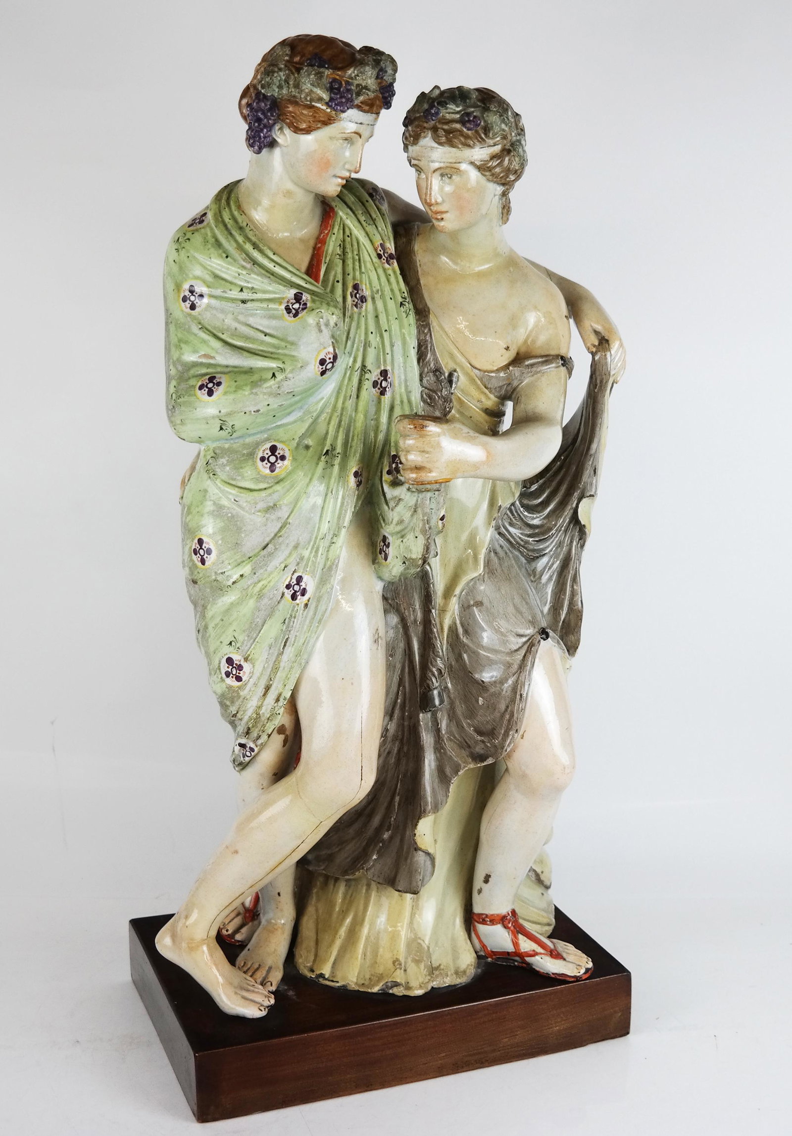 Continental Glazed Ceramic Figural Sculpture: Continental glazed ceramic figural sculpture, modeled as a Bacchanalian couple crowned with grapes and embraced around a cup of wine. [Overall: 25" H x 12" W x 10" D]. Cracks and restoration, wear and