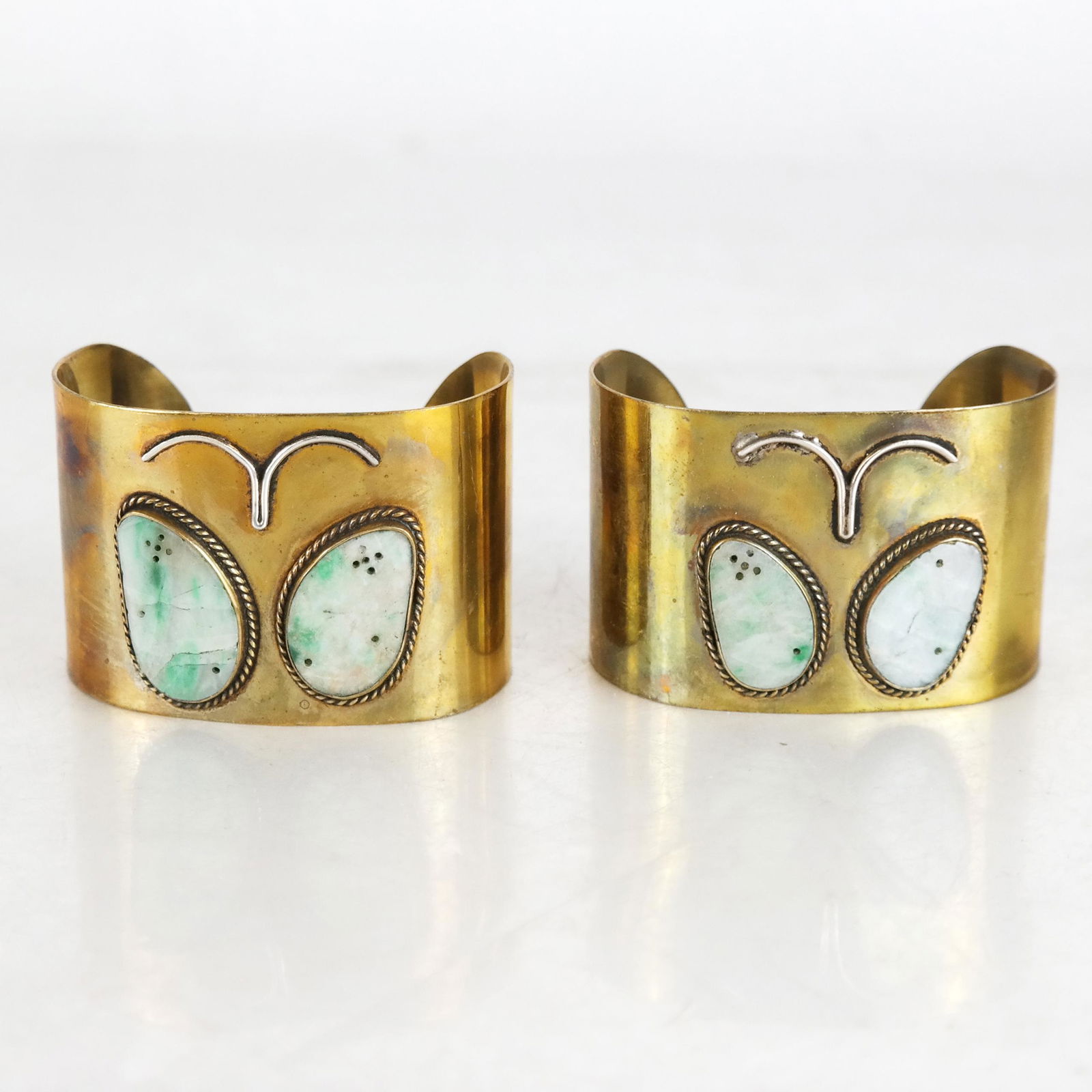Two Carved Jadite, Brass & Silver Cuff Bracelets: Two brass and silver cuff bracelets, each with two panels of jadite attached in bordered mounts, the stone hand carved with decoration. [1 3/4" H x 2 1/2" W]. In good condition.