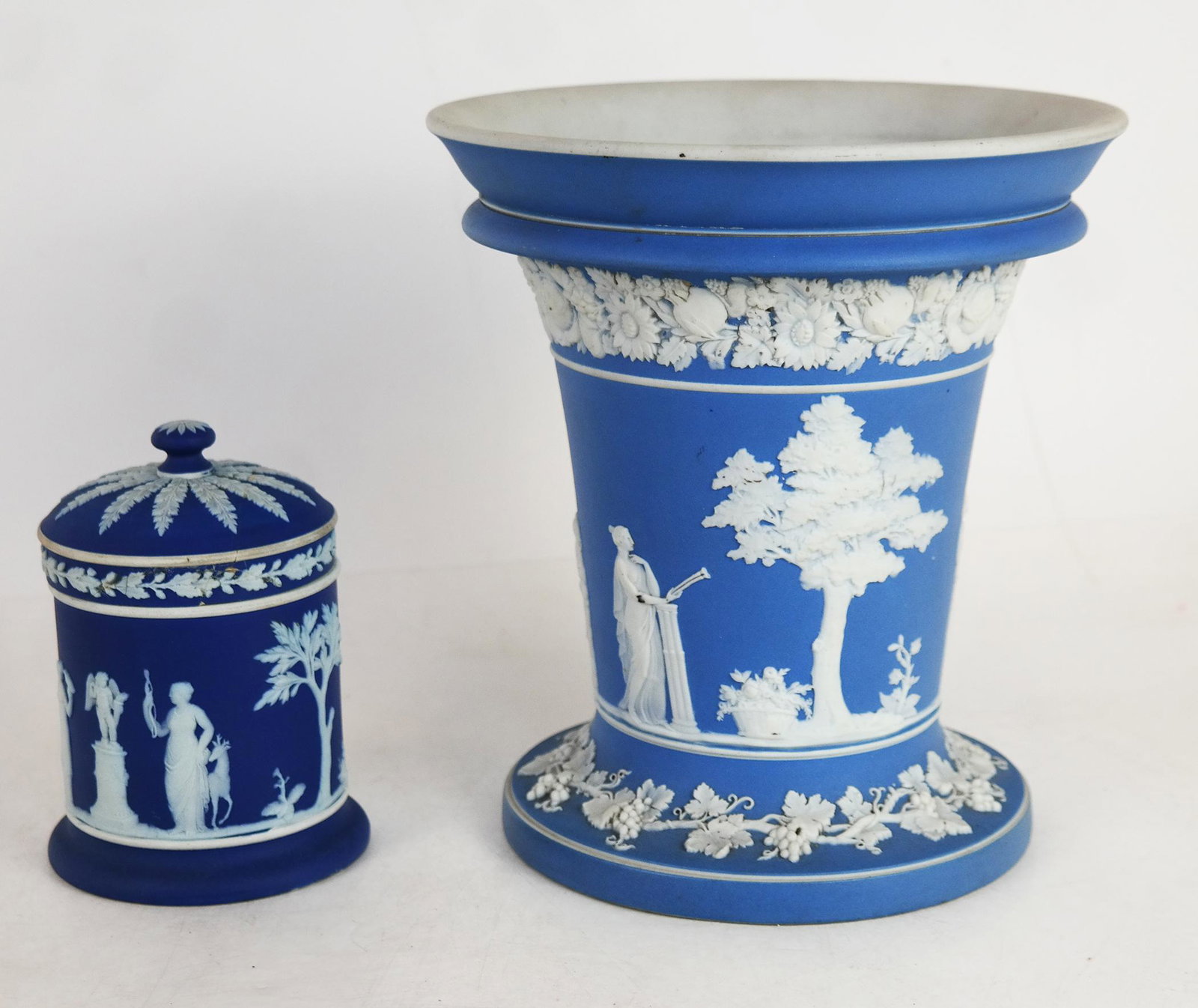 Two Wedgwood Items: Covered Jar and Vase: Wedgwood jar and cover and Neoclassical vase, the vase light blue with grape flower borders frieze, the jar and cover of darker blue, both marked. [Larger: 7 7/8" H x 7 1/2" Diam.]. Fleabite to arm of