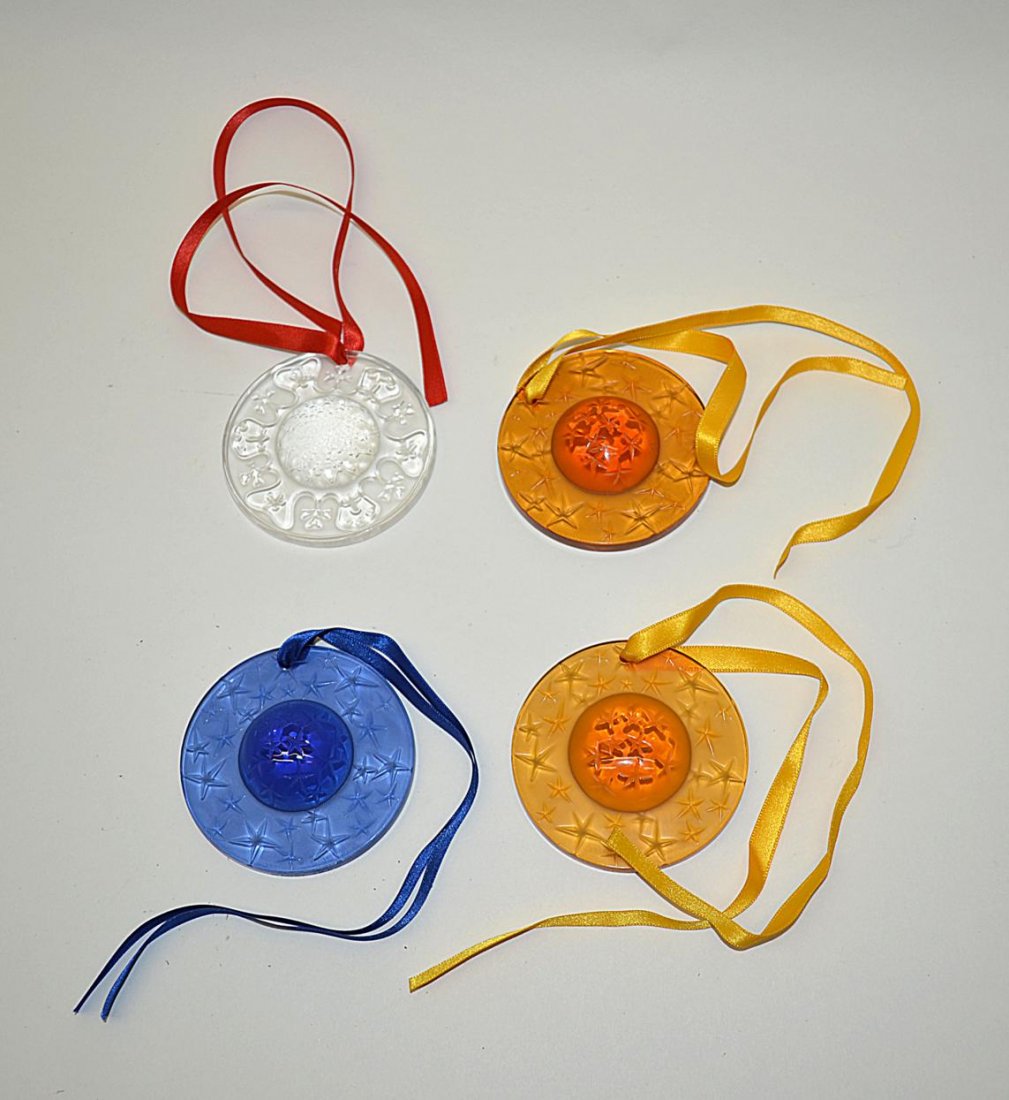 Set of 4 Lalique Holiday Ornaments: Four mould-formed circular colored crystal ornaments with raised dome center. All signed "Lalique France". 2 7/8" Dia. x 5/8" Deep. Colors include amber, blue, and clear. All in very good condition.