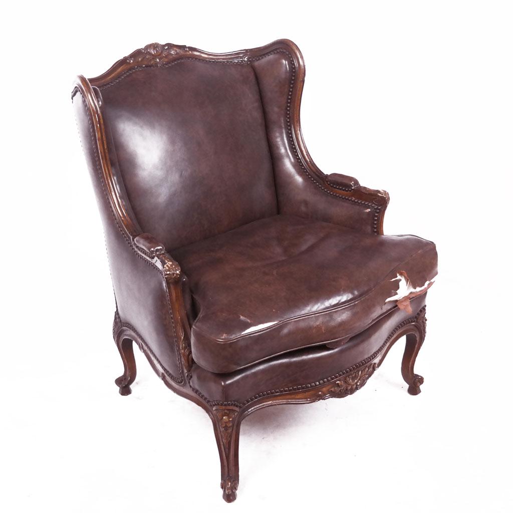 French-Style Leather Covered Bergere: French-style bergere carved with floral detail, nail head trim, with small pads to arms, covered in leather. [38 1/2" H x 30" W x 34" D]. Scuffs to wood; leather seat torn.