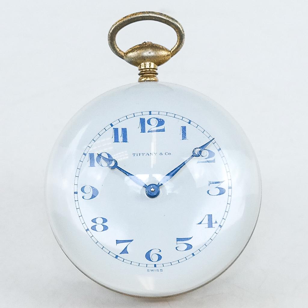 Tiffany & Co. Swiss Travel/Desk Clock: Modeled in pocket watch form with Lucite body and gilt metal band, movement stamped "Switzerland 15 Jewels" with dust and paper bag. [4 x 3 1/4 x 3 1/4 inches]. Scuffs, oxidation, wear.
