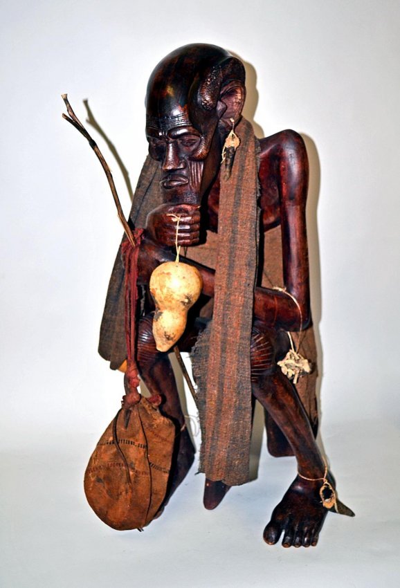 African Life-Sized Carved Wood Sculpture, Shaman: Stylized depiction of a seated African Shaman accomplished in exotic wood with detailed textures and expressive face. Accompanied by antique medicine bag, toga, dried herbs, gourd vessels, and animal 