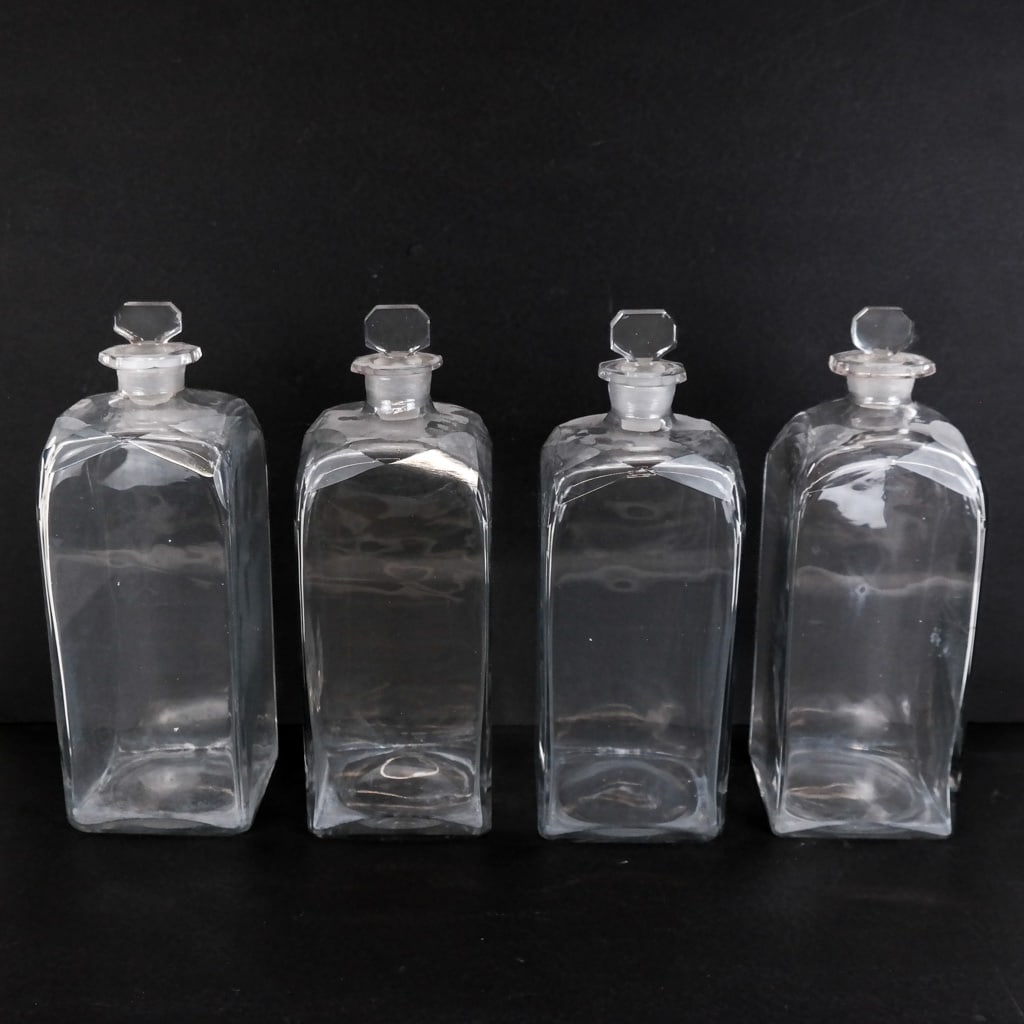 Four (4) Glass Apothecary Campaign Bottles: Four glass apothecary campaign bottles, late 19th or early 20th century, with faceted shoulder details. [11 1/2" H x 4 1/8" W x 4 1/8" D]. Chips to stoppers; residue, scuffs, marks and wear.
