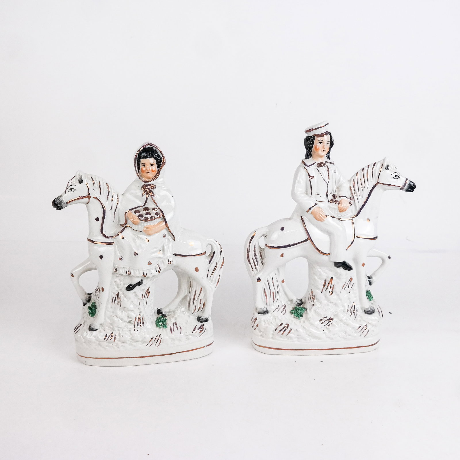 Pair of Staffordshire Riders on Horseback: Pair of Staffordshire ceramic riders on horseback, modeled as a man and woman in white with gilt trim and green flora. [9" H x 6 1/2" W x 3" D]. One horse with repaired tail.
