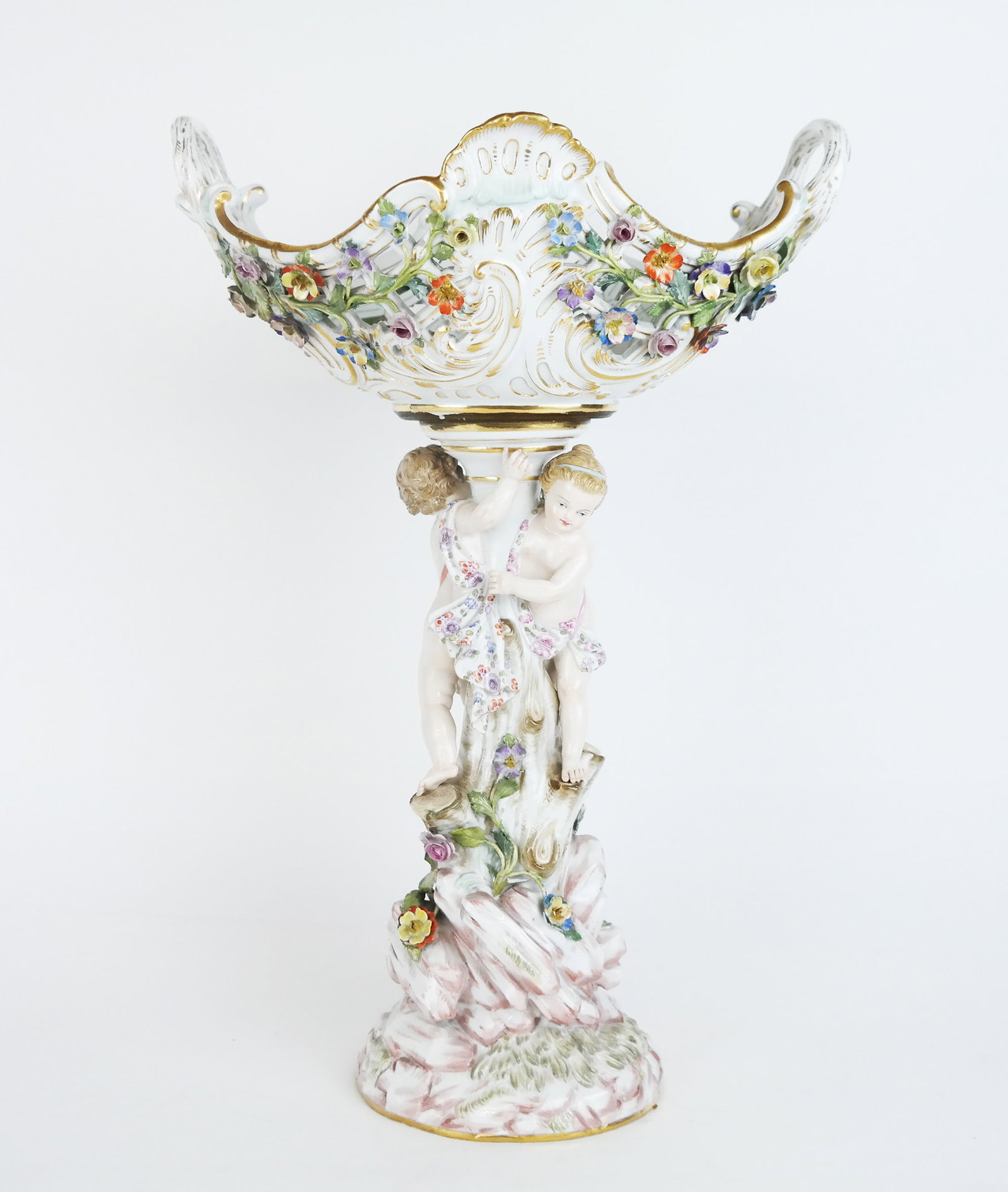 Meissen Tall Figural Centerpiece: Meissen tall figural centerpiece comprising an open reticulated bowl on a putti-mounted figural and rock-form base post. [16" H x 11" W x 8" D]. Small chips to small mounted flowers.