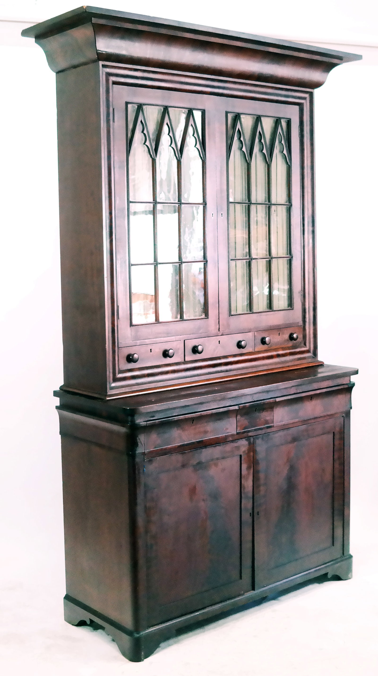 Gothic Revival Flame Mahogany Cabinet: Gothic Revival flame mahogany cabinet, 19th century, in two parts, with Gothic mullion glass doors, two drawers and dual door below. [95" H x 50" W x 20" D]. Wear commensurate with age.