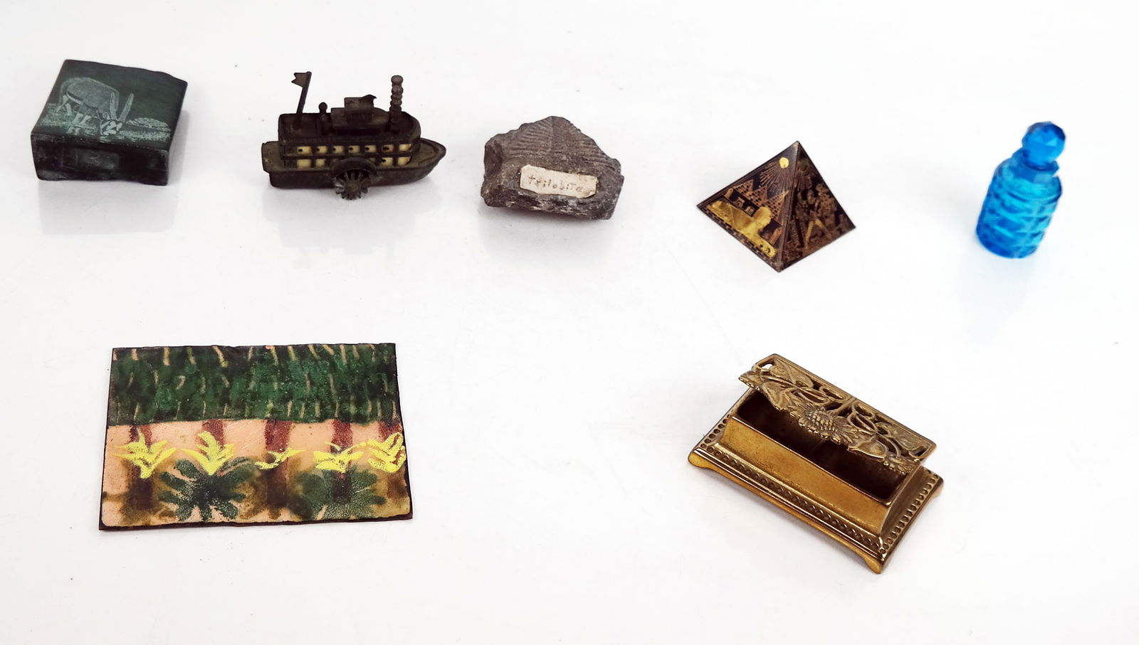 Seven Small Decorative Objects: Group of small decorative and desktop objects, including an etched brass pyramid paperweight; trilobite fossil; paddleboat pencil sharpener; enameled copper tile; brass stamp holder; etched South Afri