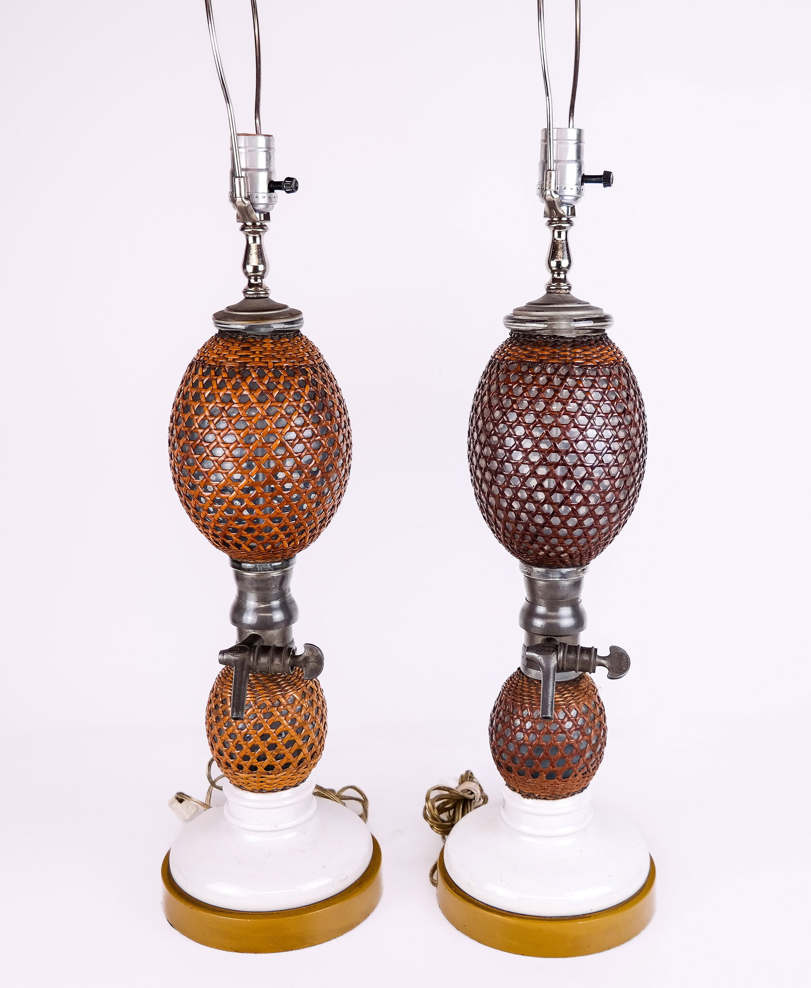 Pair of Unusual Spigot & Seltzer Lamps: Pair of double-tier glass and metal spigot seltzers wrapped in cane on porcelain and wood bases, mounted as a pair of lamps. [33" H x 7" diameter]. In good condition. Condition reports and requests fo