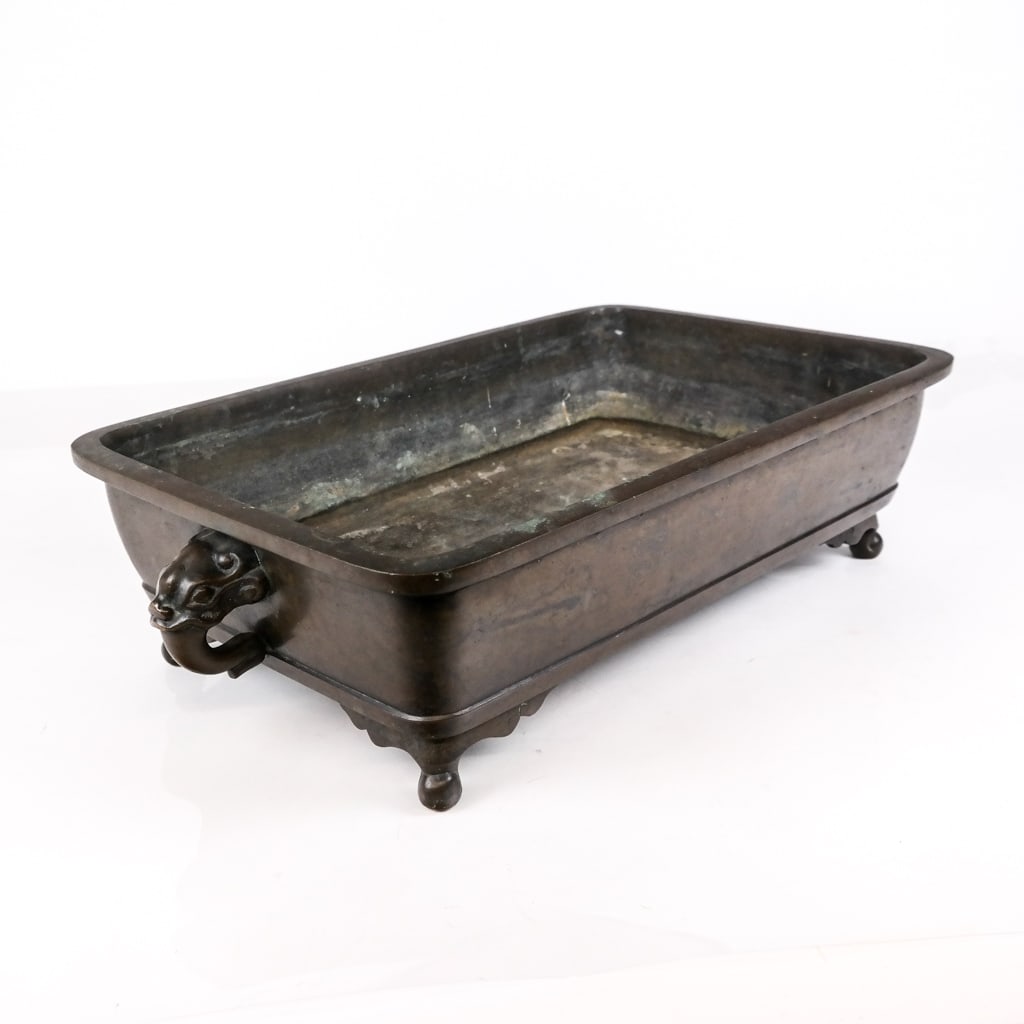 Japanese Bronze Suiban Jardiniere: Japanese bronze suiban or jardiniere, 20th century, with two mask form handles, on ball feet. [5" H 20.5" W x 12" D]. Overall condition is good with scattered wear to patina.