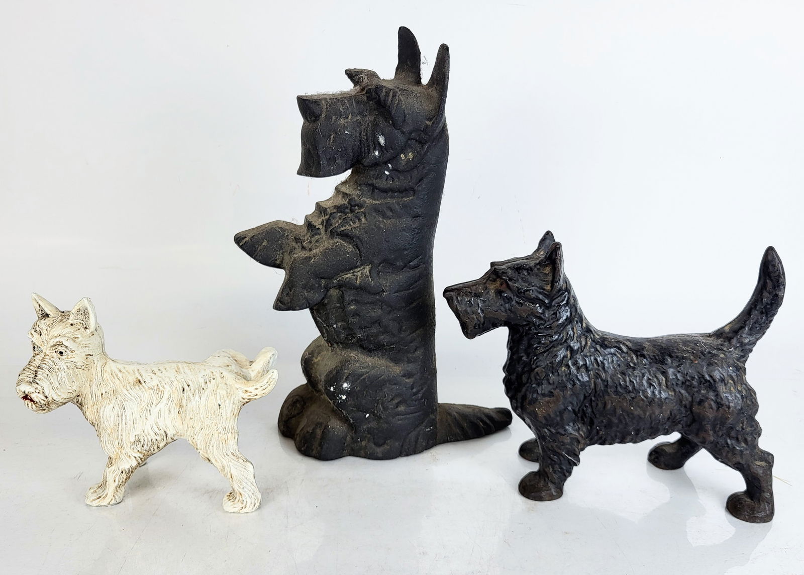 Cast Iron Terrier Doorstops, 3: Three cast iron terrier or scottie form doorstops, comprising an American cast iron of a scottie, a white-painted cast iron of a terrier with its rear leg up, and a cast iron terrier on its hind legs.