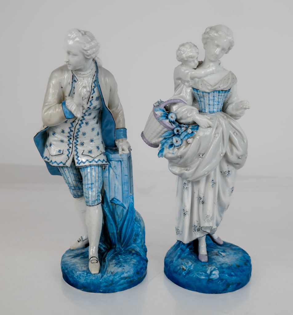 Porcelain Blue and White Figures, 2: Pair of porcelain blue and white figures, likely German, with both figures dressed in 18th century apparel and one figure leaning up against a pediment while the other carries a baby and a basket of f