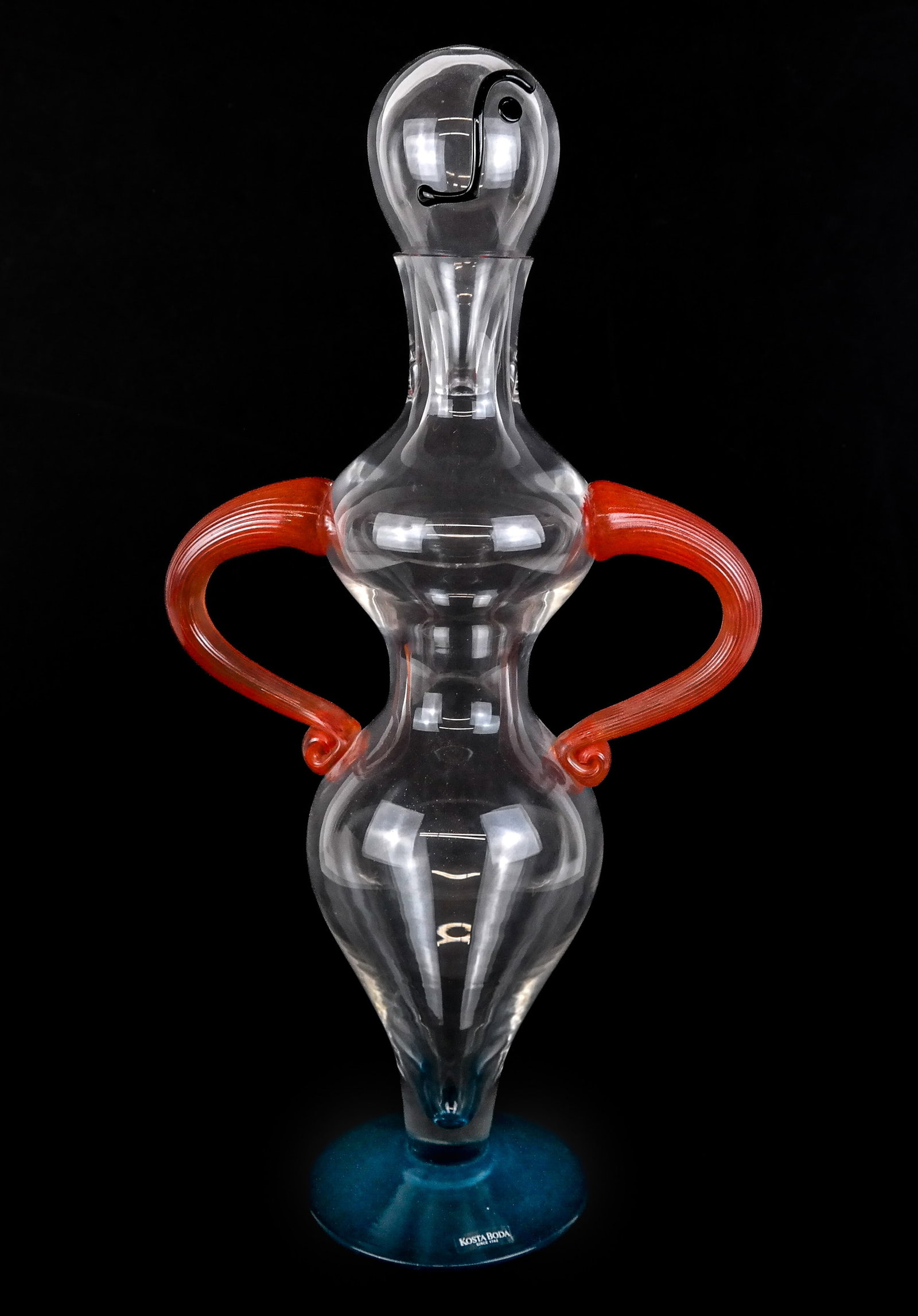 Kjell Engman for Kosta Boda "Fenix" Decanter: Kjell Engman for Kosta Boda blown glass decanter, Swedish, 20th century, with black glass accents to face, a clear glass body, orange reticulated glass arms, a blue glass base, signed Kosta Boda K. En