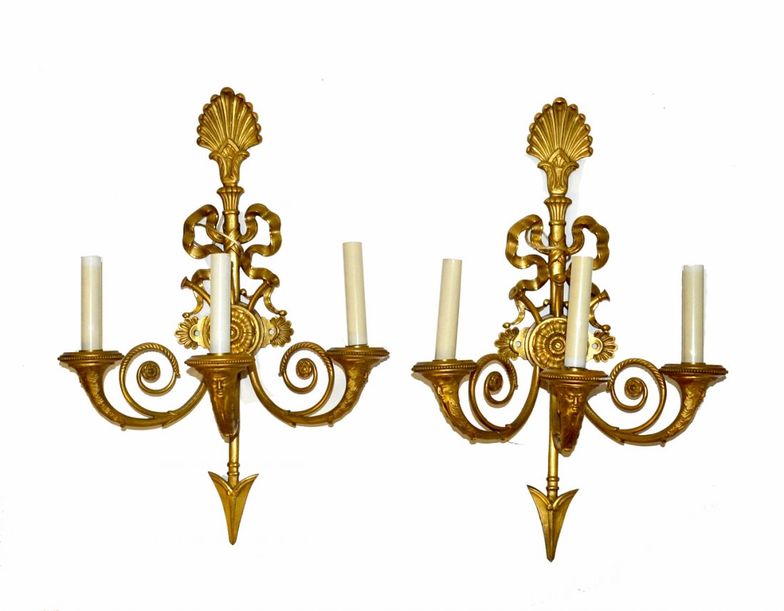 Pair of Bronze Neo-Classical Three Arm Sconces: Each feature arrow and ribbon-form backplates with acanthus finials, scrolling tendril accents, and grotesque face details on each arm. [14"W x 8"D x 22"H]