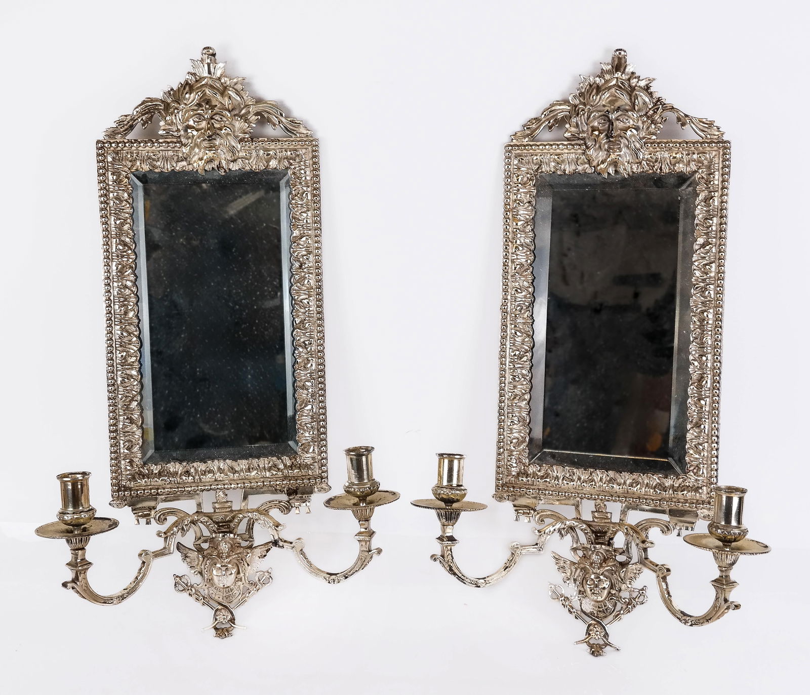 Pair of Silvered Metal Two Light Sconces: Pair of silvered metal and beveled glass two light sconces, accented with masks. [23.5" H x 13" W x 5.25" D]. Overall condition is good, with some losses to metal and spots of tarnishing. CABINET 2