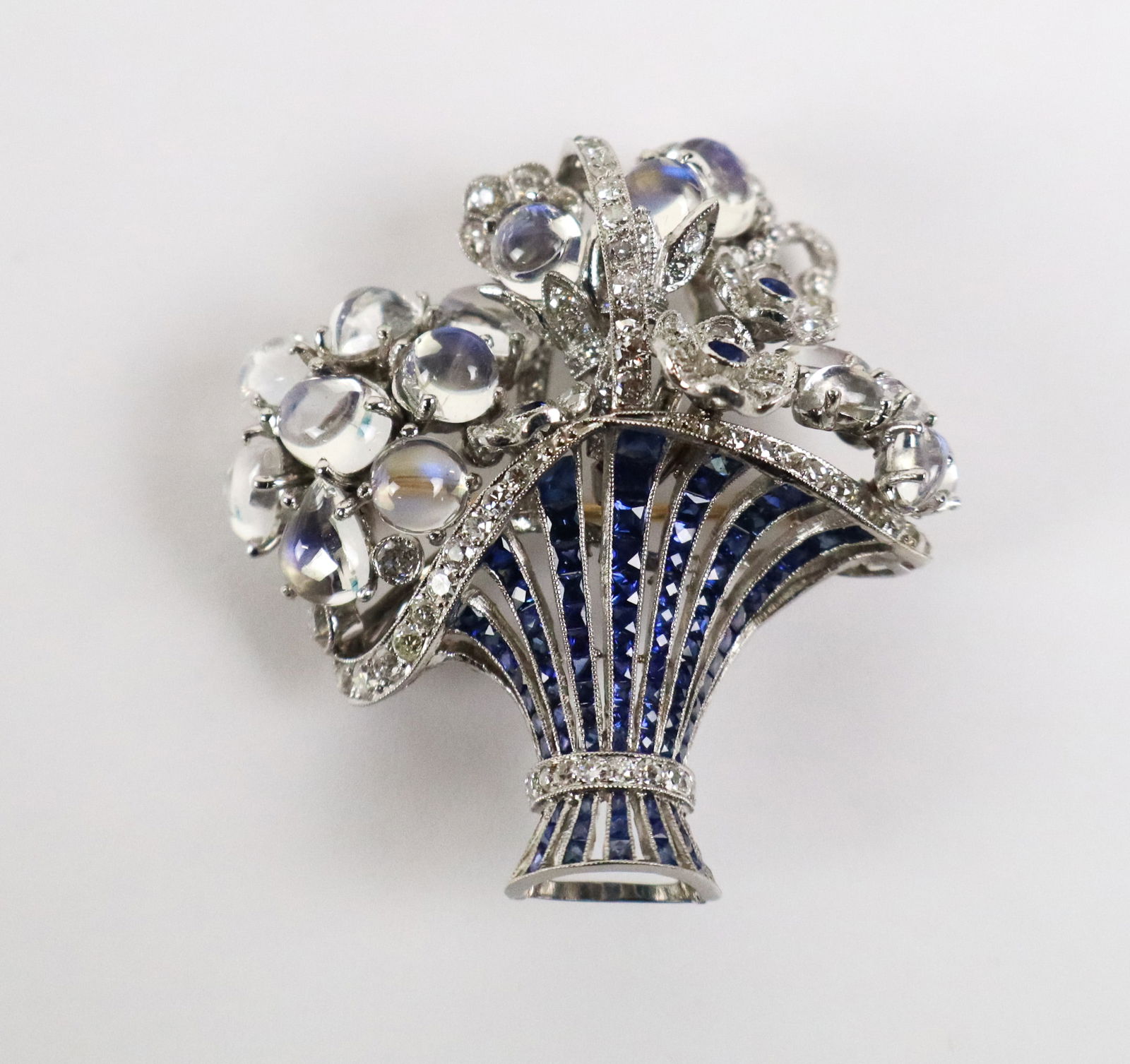 Fred Leighton Moonstone, Diamond & Sapphire Brooch: Fred Leighton cabochon moonstone, diamond and sapphire flower in a basket brooch, the basket defined by graduating bands of French-cut sapphires and circular diamond border and handle, signed on the y