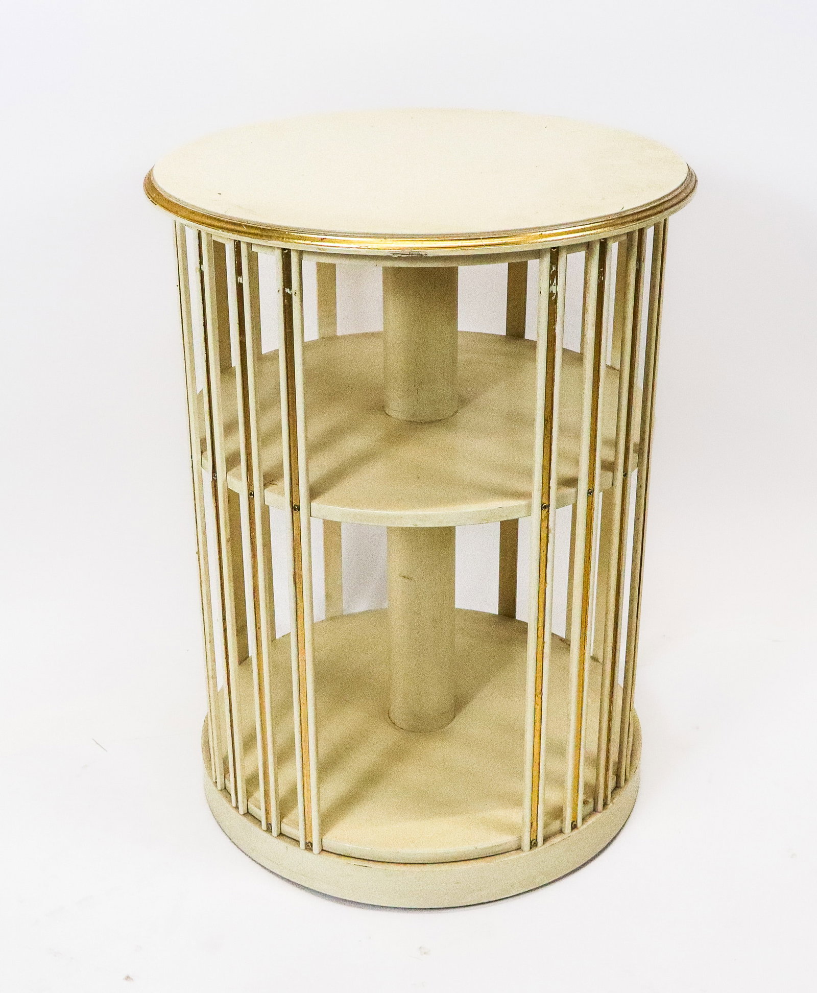 Modern Painted Round Book Stand: Modern painted round book stand in 3 tiers, with off white paint and gilt trim, revolving on wheels. [26" H x 19" diameter]. Slight chips and discoloring.