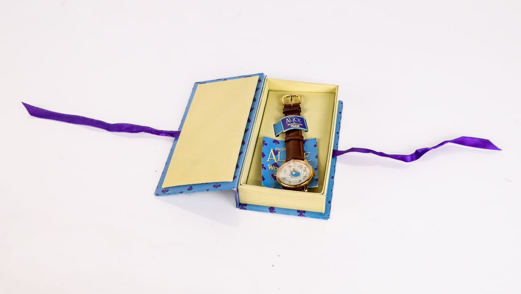 Alice in Wonderland 45th Anniversary Watch: Alice and Wonderland 45th anniversary watch in its original box, with plastic wrap around watch face and back. [1.25" Dial, .25" Thickness, 7" Strap Length] Overall condition is good, with the watch n