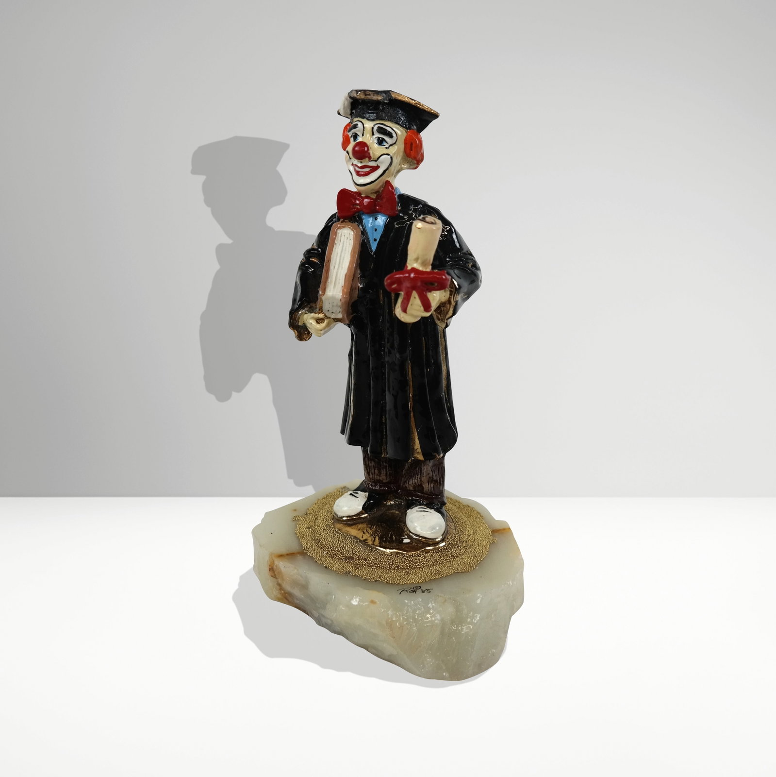 Ron Lee Clown Graduate Sculpture: Ron Lee (American 20th century) sculpture of a graduated clown, 1985, signed Ron to onyx base. [9" H x 5" W x 4" D]. Condition is good with a repair to cap.