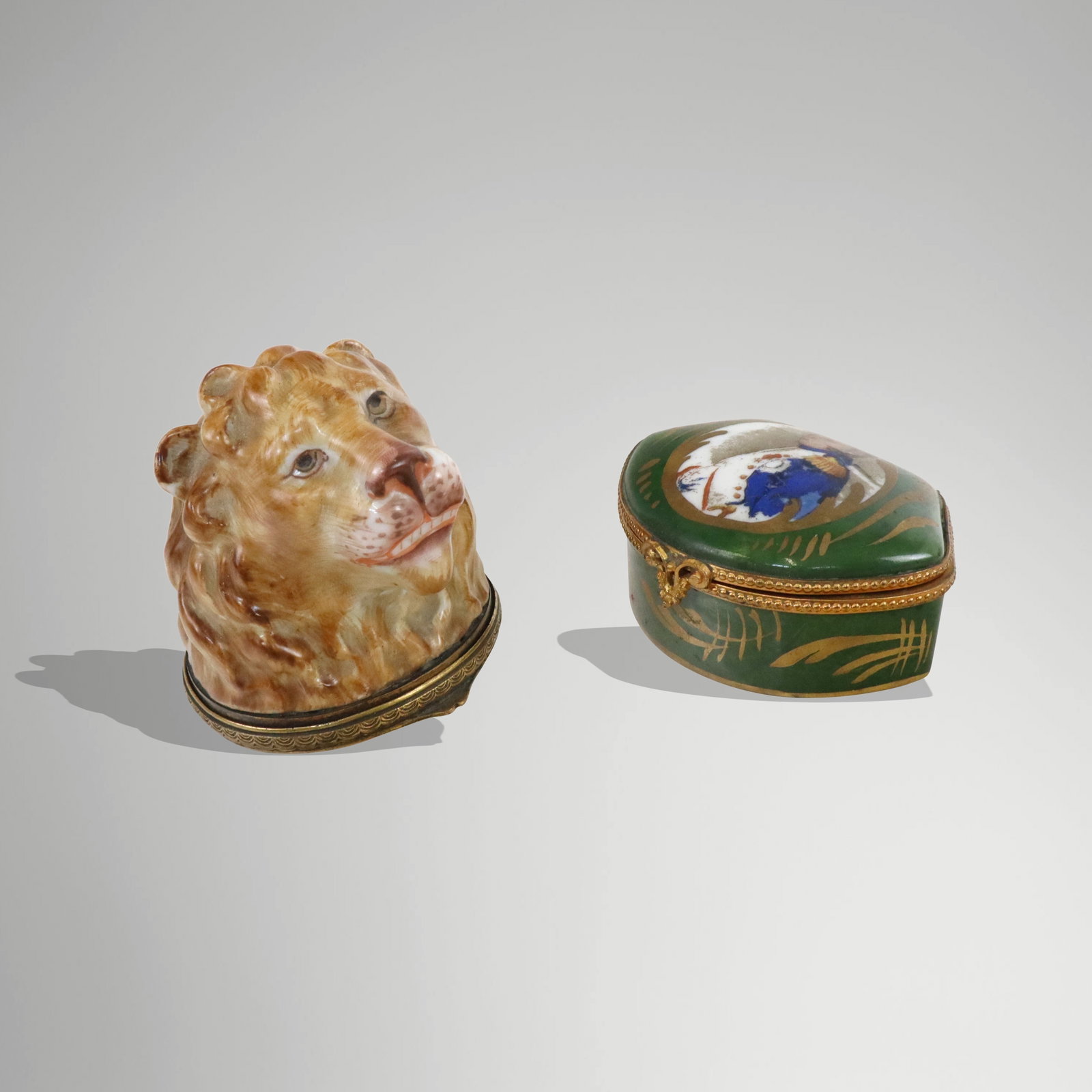 Two Porcelain Decorative Small Boxes: Two porcelain decorative small boxes, early 20th century, comprising an English lions head with brass hinged bottom having engraved designs [2.5" H]; and a Limoges style Napoleon top green painted