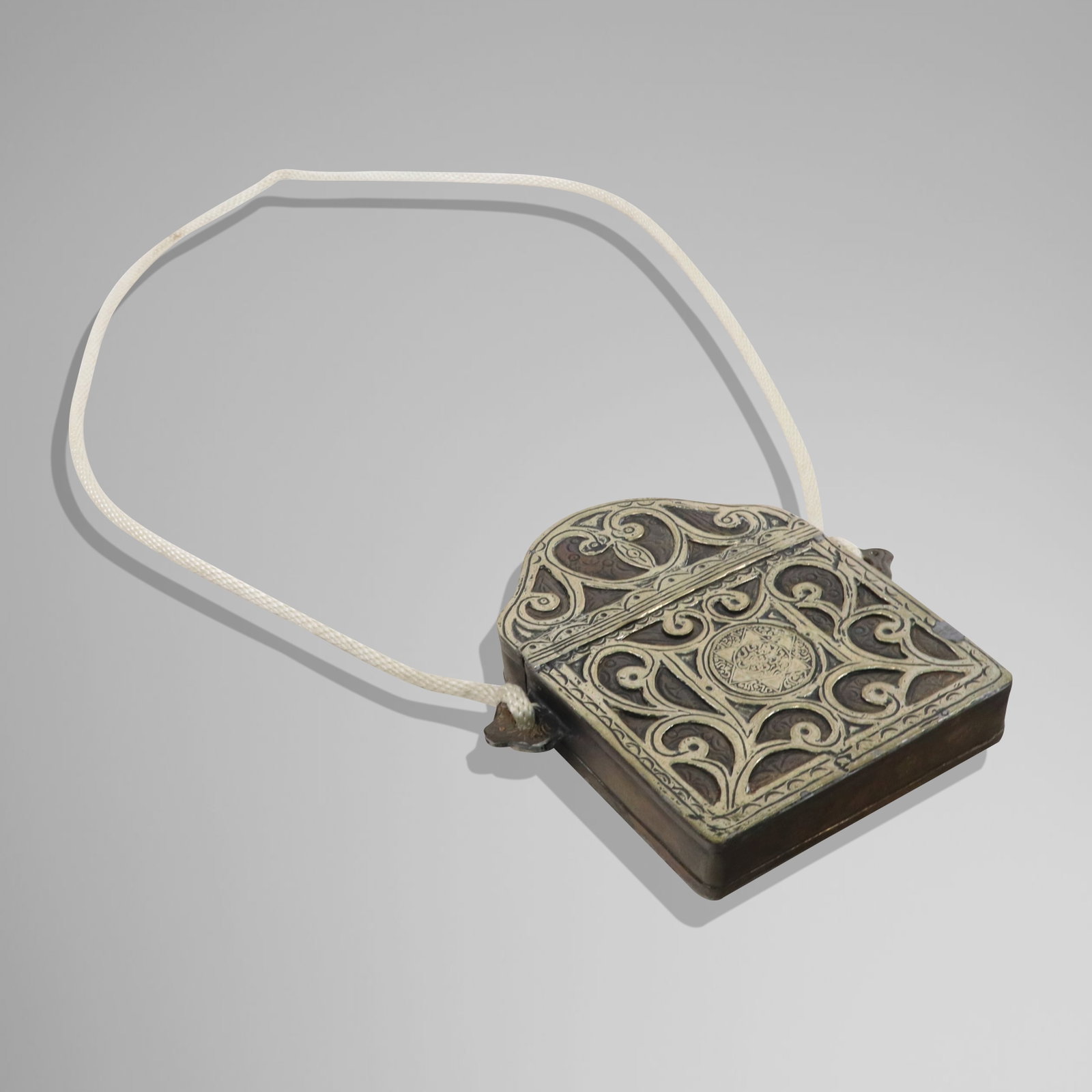 Islamic Metal Box on Necklace: Islamic metal box; hinged top box with brushed silvered metal and unpolished, with engraved designs, tied on a rope necklace. [6" x 6"]. Aged dents and spot repairs.