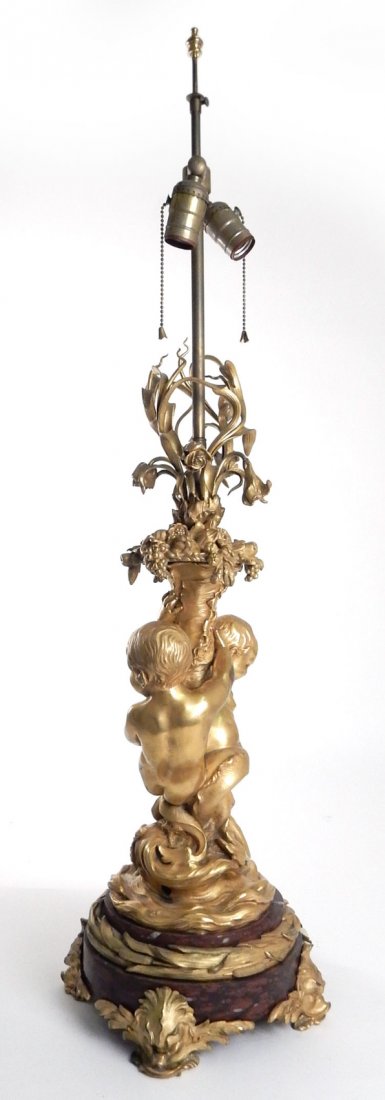 Gilded Bronze Ormolu Figural Lamp: Ormolu gilded bronze figural allegorical lamp featuring two children with mermaid tails entwined about a cornucopia on a marble base with ormolu dolphin feet. [26" H x 38 1/2" base to finial x 10"]. I