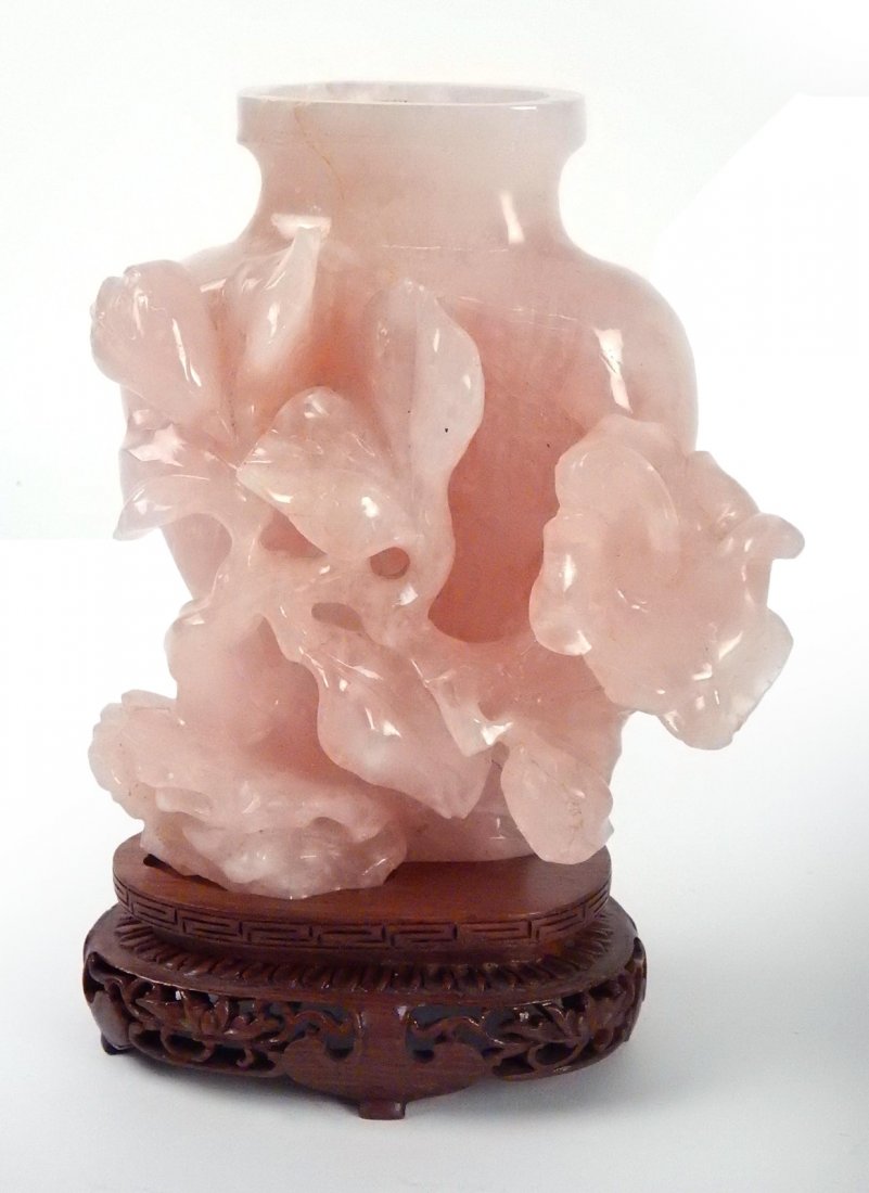 Carved Rose Quartz Vase: Carved rose quartz urn/vase with leafy branch and wood base. [5 1/2" H x 5 1/4" W; 6 5/8" with wood stand]. Fractures, missing lid.