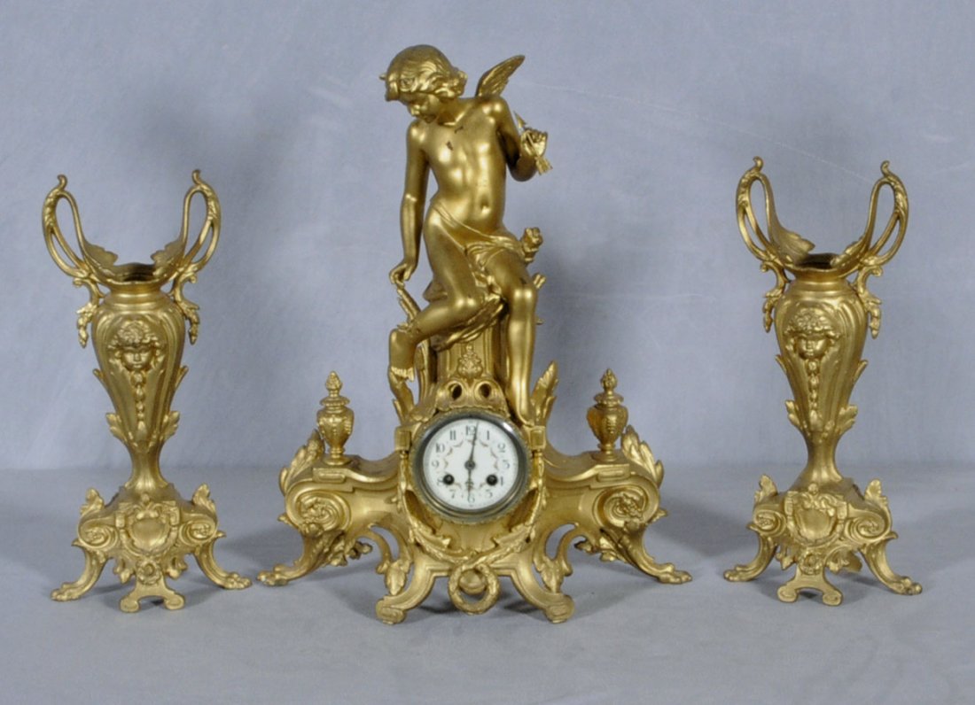 3 PC. GILT SPELTER CLOCK SET. CONSISTING OF AN ANGE: 3 PC. GILT SPELTER CLOCK SET. CONSISTING OF AN ANGEL SITTING ATOP THE CLOCK. CLOCK HAS A SHAPED BASE. PAIR OF FACIAL RELIEF DESIGN URNS. 19 3/4" H X 16" W -CLOCK. URNS 15 1/2" H X 7 1/4" W.
