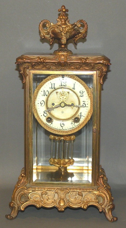 Ansonia “Crown” crystal regulator clock: ca. 1910; eight day time and strike spring driven movement with visible escapement and ornate pendulum in a brass case with urn top and porcelain dial, 15 ½”x 8”;