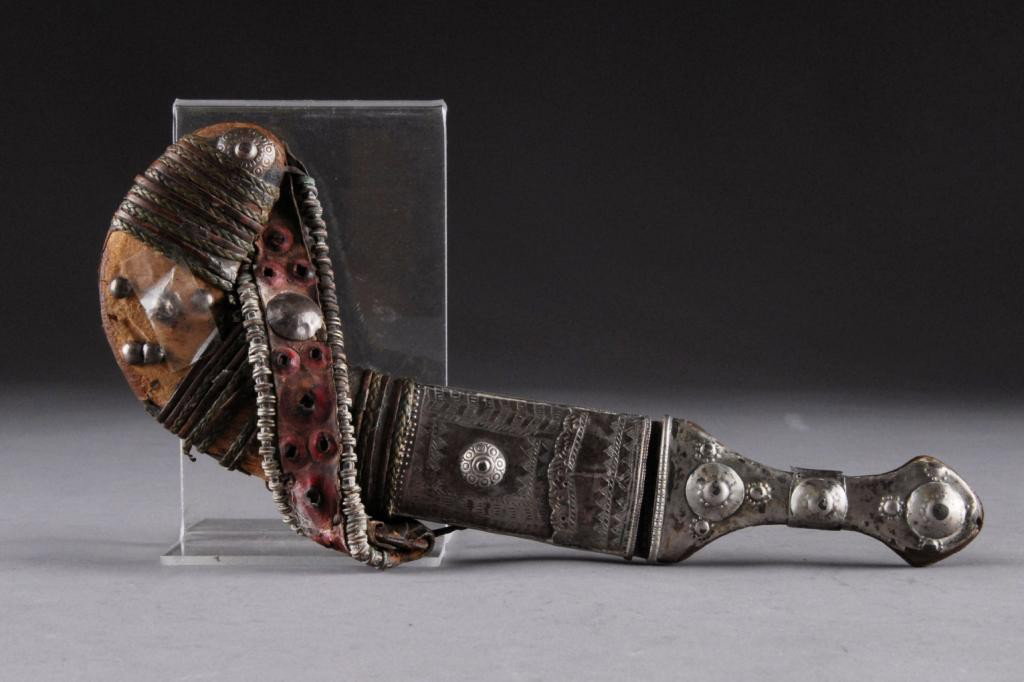 AN ARABIAN KNIFE,: the shaped blade with foliate engraving, the handle with carved horn and silver strap to the pommel, the sheath of leather and silver. Length: 11"