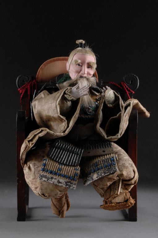 AN ANTIQUE CHINESE WARRIOR DOLL FIGURE,: modeled in a seated position wearing silk robes with parcel gilt and metal mounted armor, set in a carved wood throne chair with good luck symbol detailed sides. Height: 16"