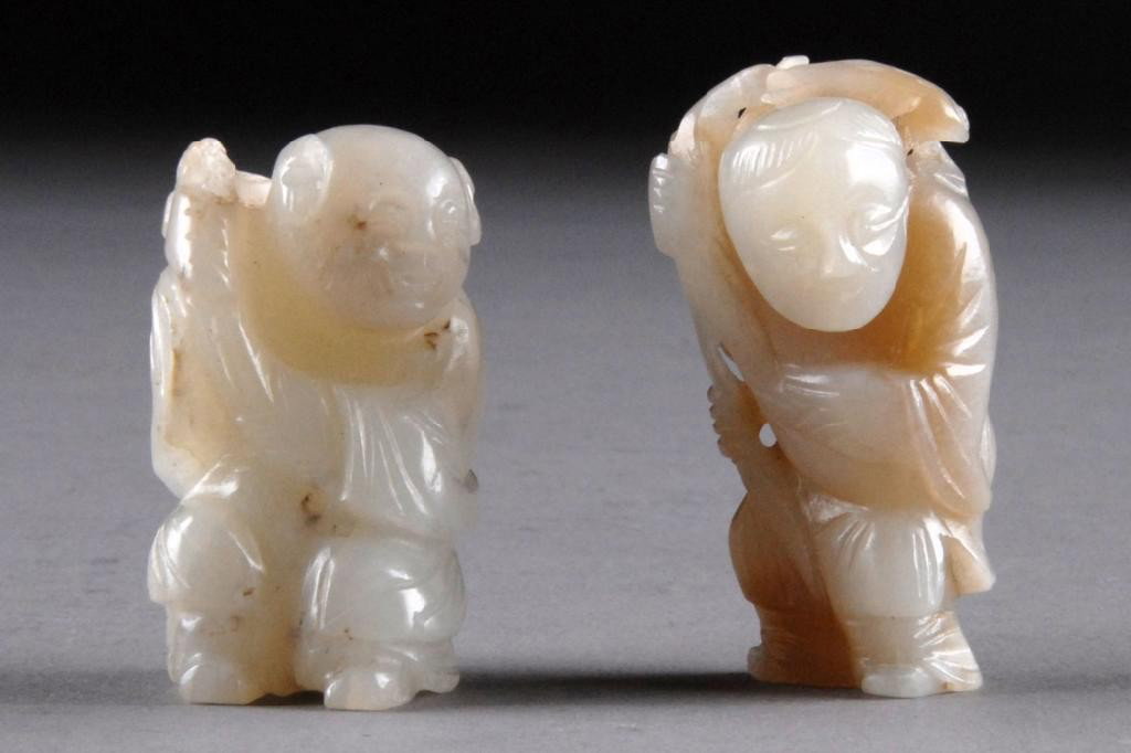 TWO CHINESE WHITE JADE FIGURAL PENDANTS,: possibly modeled as the smiling twins Hehe Erxian with each having a bundle of ruyi and long stemmed lotus bundles hoisted over right shoulder and wearing flowing robes with a smiling expression, with