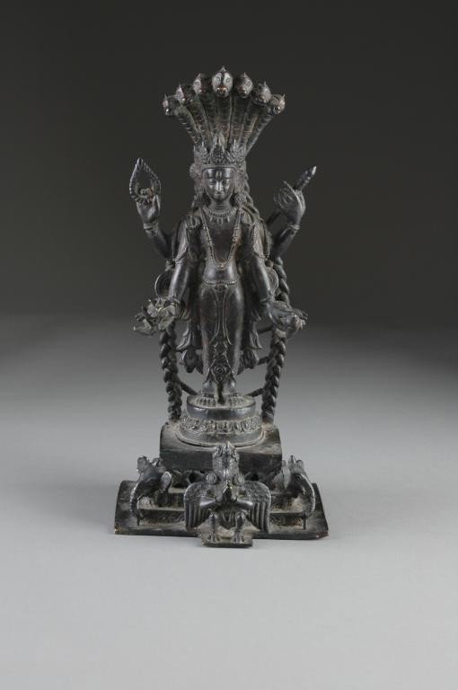 AN INDONESIAN BRONZE SCULPTURE OF BUDDHA,: shown with four arms, standing holding a lotus, scepter, flame, and vial, wearing elaborate headdress and ear pendants and multiple necklaces, standing on a raised pedestal, on a stepped plinth. Heigh
