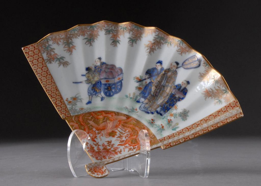 TWO ANTIQUE IMARI PARCEL GILT AND ENAMELED DISHES,: LATE MEIJI PERIOD, one of fan form and centering an elder with attendants watching two youths pulling a loaded cart amidst blossoming foliage beneath a rim of delicate polychrome painted andgilt bambo