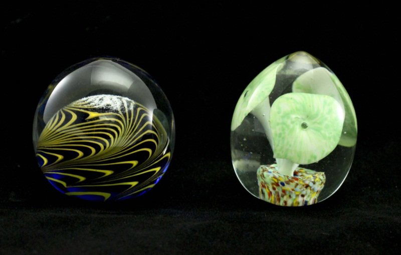 (2) Modern Art Glass Paperweights: Lot of (2) art glass paperweights. Egg-shaped paperweight with green Calla lilies inside (measures 3"d x 3.25". Dark blue paperweight has yellow caning encased in clear glass (measures 3" x 3.25"). Me