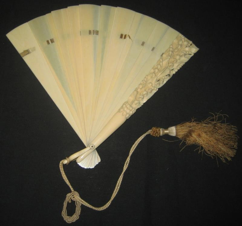 ANTIQUE CARVED IVORY FOLDING HAND FAN: Composed of sixteen plain tapered ribs connected by woven silk ribbon (imp.) and two guardsticks, one finely carved with lily-of-the-valley spray in high relief, 19th century. 9 1/2 x 15 1/2 in (open)