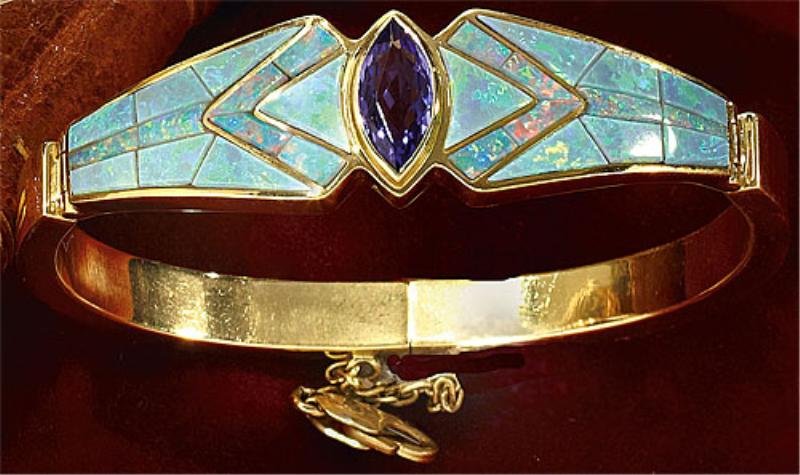 TANZANITE AND OPAL BRACELET, DAVID FREELAND: 18k yellow gold oval hinged bangle centering a marquise cut Tanzanite measuring 12.5mm x 6mm. Center stone is flanked by invisible set opals in a mosaic design. Top width is 16mm, inside bangle dimens