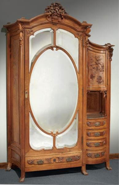 ANTIQUE CONTINENTAL ART NOUVEAU WALNUT ARMOIRE: With elaborate carved floral crest over the arched full length mirrored door of shaped beveled panels, beside a convex bird and floral carved panel door, an open display area and four short drawers, c
