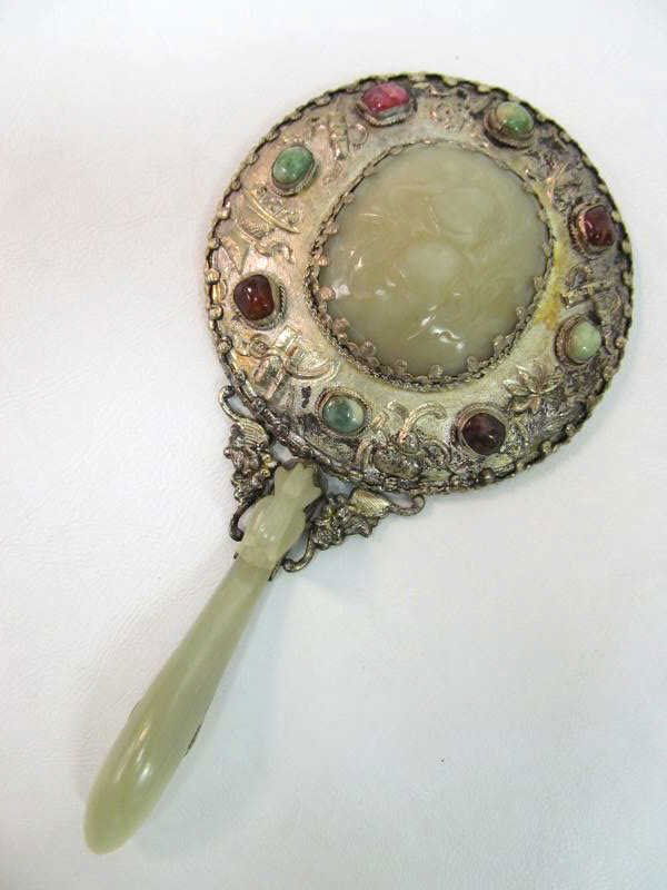 CHINESE SILVER AND WHITE JADE HAND MIRROR: Silver on copper mirror inset with peach motifs and carved jade plaque in the center surrounded by various mineral cabochons, the handle in the form of a dragon buckle, 18th/19th century. Length: 8 in