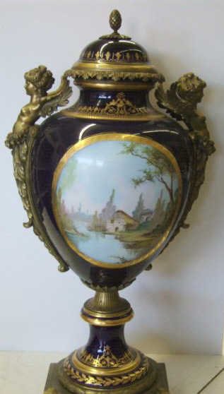 SEVRES BRONZE MOUNTED PORCELAIN URN WITH LID: The cobalt blue ovoid body painted with two reserves, one depicting romantic scene, the other river landscape within elaborate gilding, indistinctly signed, mounted at top winged figural and foliate h